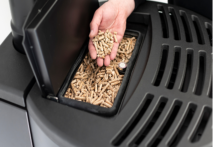 Eine Hand hält Holzpellets über ein offenes Fach, das mit weiteren Pellets in einem Pelletofen gefüllt ist, und zeigt, dass Brennstoff nachgefüllt wird.