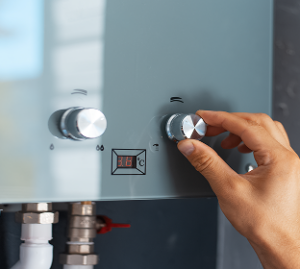 Eine Hand dreht den Temperaturregler eines modernen tanklosen Warmwasserbereiters, dessen Digitalanzeige 38 °C anzeigt. Unter dem Gerät sind Rohre und Anschlüsse zu sehen.