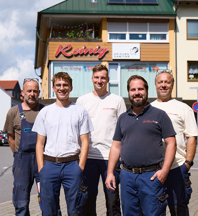 Fünf Männer in Arbeitsuniformen stehen lächelnd vor einem Gebäude mit der Aufschrift Reinig. Das Gebäude ist gelb mit grüner Umrandung, und die Gruppe steht auf einer gepflasterten Straße unter einem teilweise bewölkten Himmel.