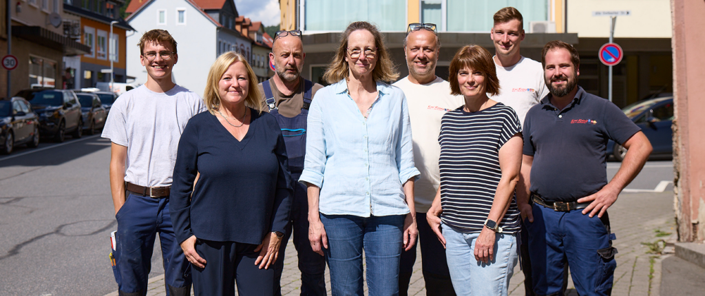 Eine Gruppe von acht Personen, fünf Männer und drei Frauen, steht lächelnd auf einer sonnigen Straße mit Gebäuden und geparkten Autos im Hintergrund zusammen.