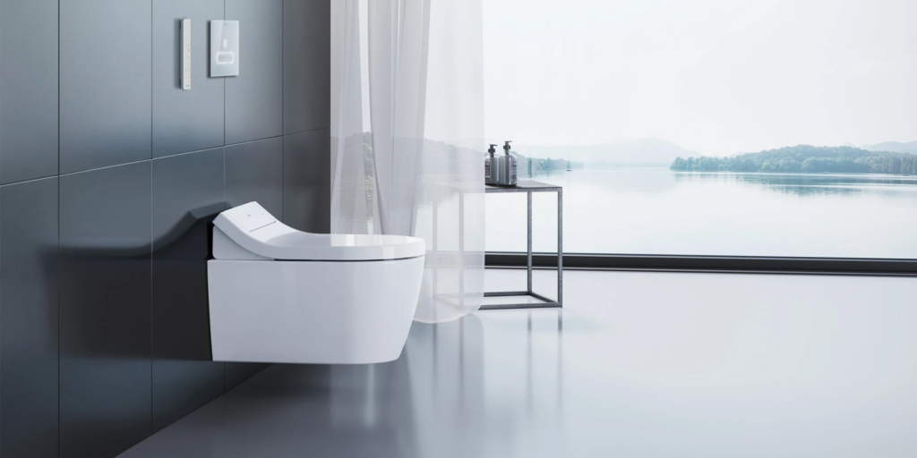 Eine moderne, an der Wand montierte, intelligente Toilette in einem minimalistischen Badezimmer mit dunklen Fliesen, einem weißen Vorhang, einem kleinen Tisch mit Seifenspendern und großen Fenstern mit Blick auf einen ruhigen Seeblick.