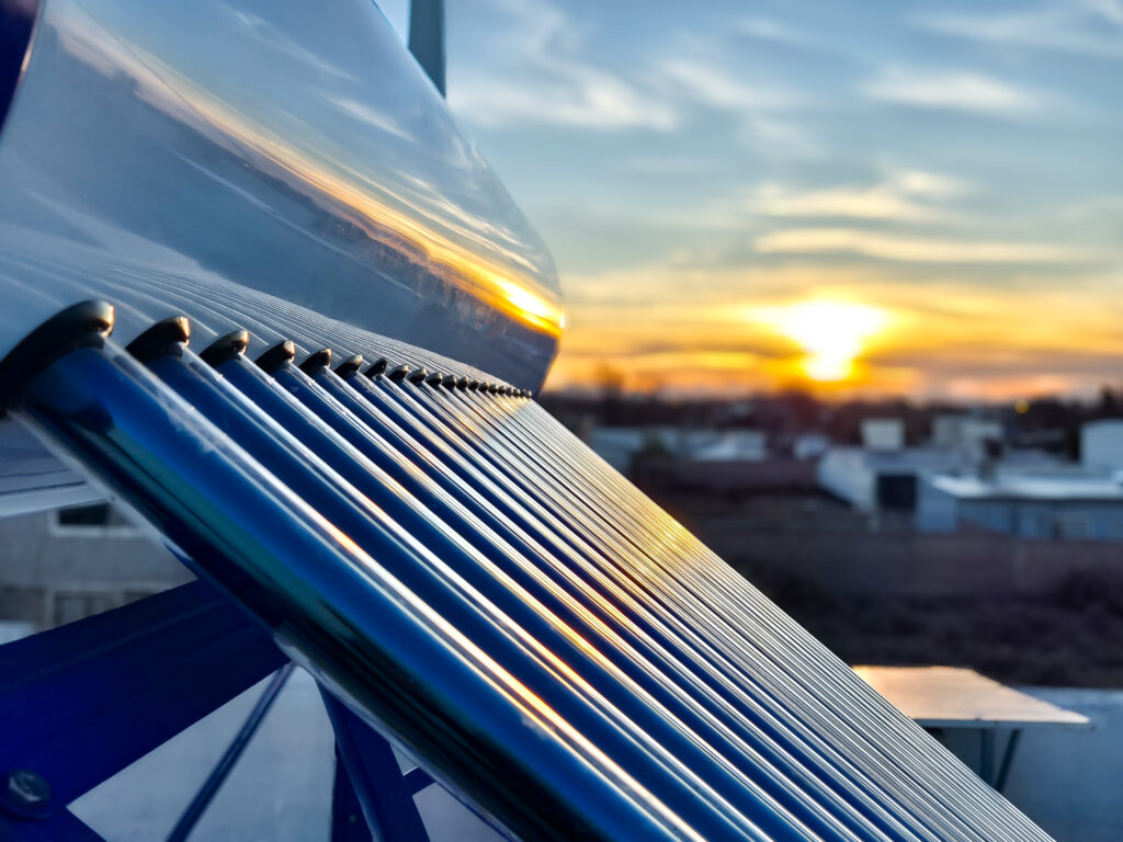 Nahaufnahme von Röhren eines Solarboilers auf einem Dach, die das Sonnenlicht reflektieren, mit einer Stadtlandschaft und einem leuchtenden Sonnenuntergang im Hintergrund.