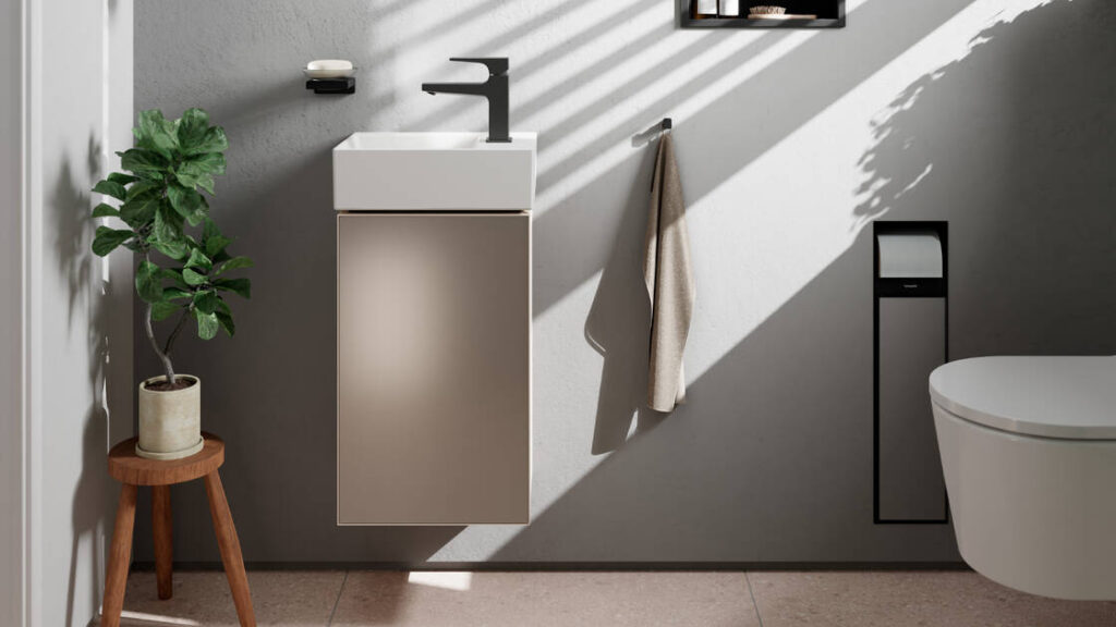 Minimalistisches Badezimmer mit einem kleinen Waschbecken und einem schwarzen Wasserhahn, einem beigen Schrank, einem an der Wand hängenden Handtuch, einer Topfpflanze auf einem Holzhocker, einer Toilette und Sonnenlicht, das durch Lamellen fällt und Schatten an die Wand wirft.